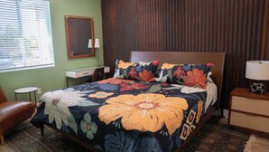 Une chambre à coucher avec une couvre-lit à motifs floraux, une tête de lit en bois, une table de chevet avec une lampe, une chaise et une fenêtre avec des stores.