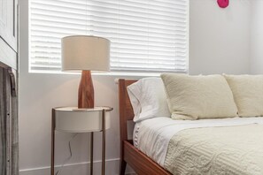 A bedroom with a wooden bed, white bedding, a round table with a lamp, and a window with blinds.