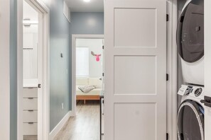 A laundry room with a washing machine, a bed with a white headboard, and a hallway leading to a room with a window and blinds.