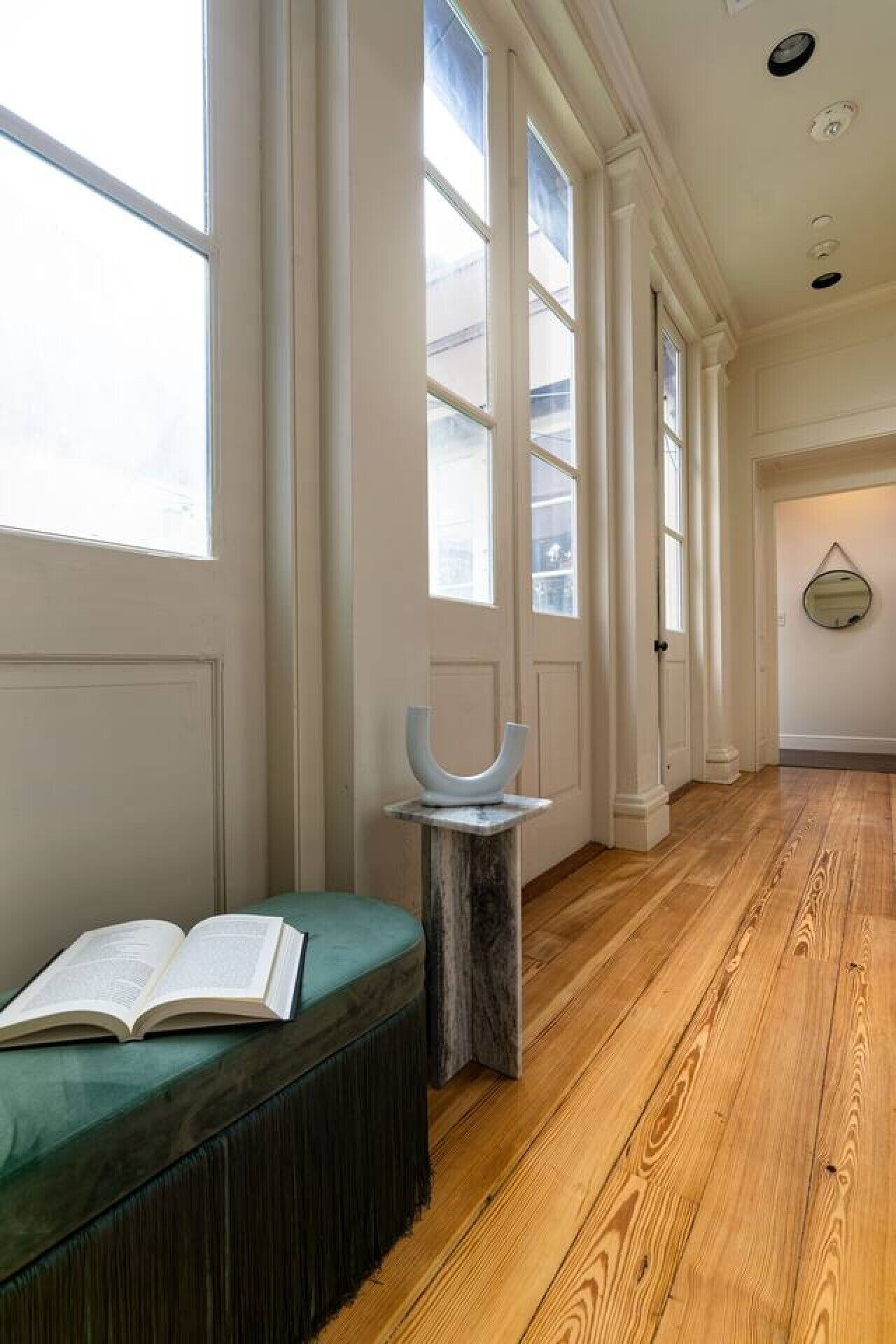 A hallway with a wooden floor, a green upholstered bench, and a marble pedestal with a sculpture.