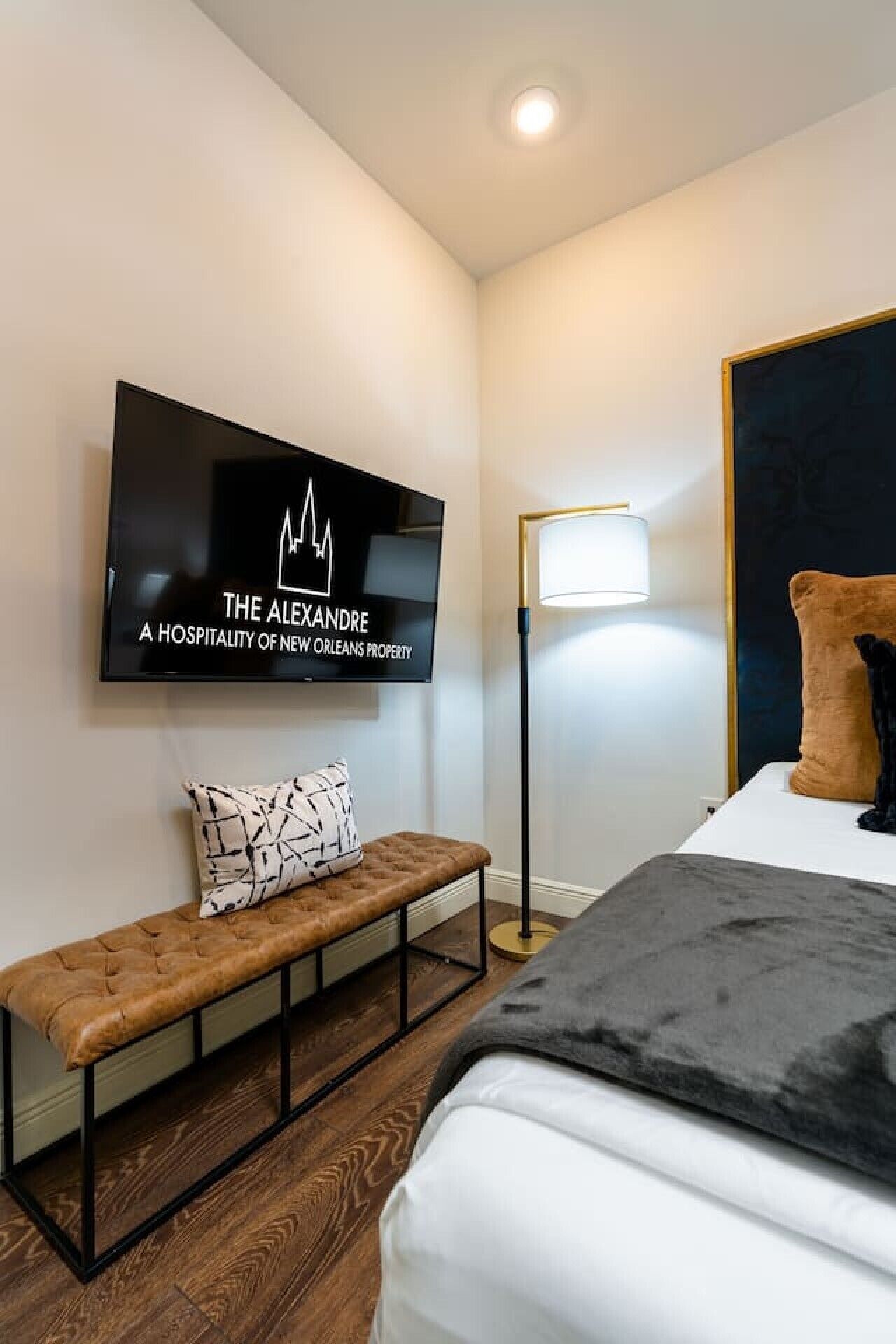 A hotel room with a bed, a bench, a TV mounted on the wall, and a floor lamp.
