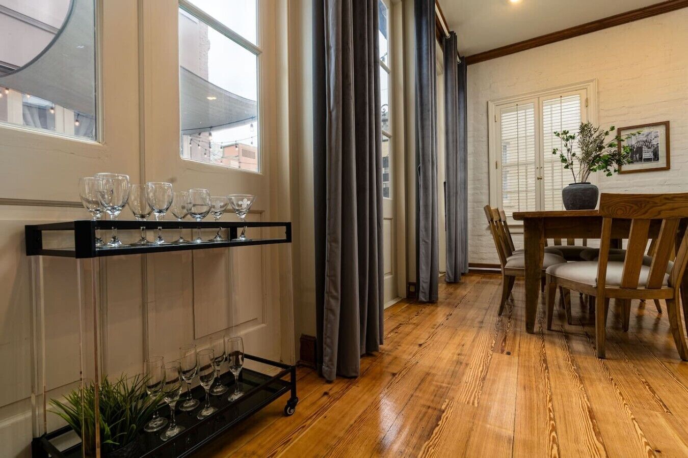 Una habitación con piso de madera, una mesa de comedor con sillas, una repisa negra con copas de vino y una ventana con cortinas.