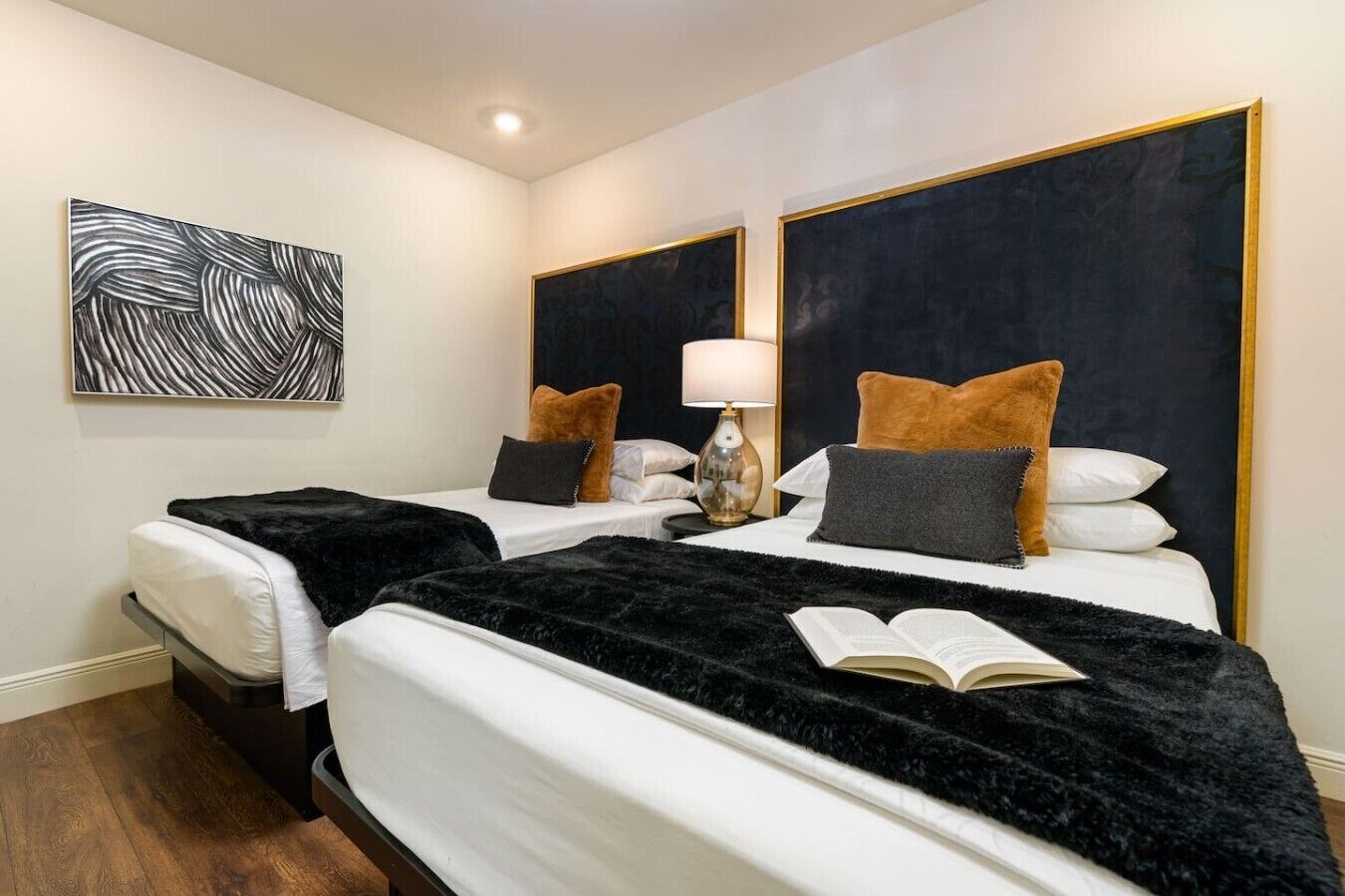 Una habitación de hotel con dos camas, un cuadro abstracto en blanco y negro, una mesita de noche con una lámpara y un libro sobre la cama izquierda.
