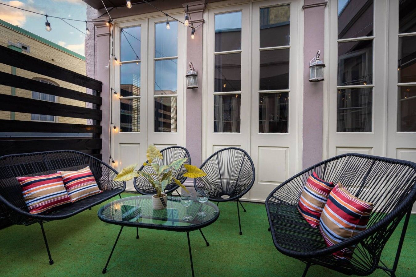 An outdoor seating area with wicker chairs and a glass table, surrounded by a green carpet and a building with large windows.