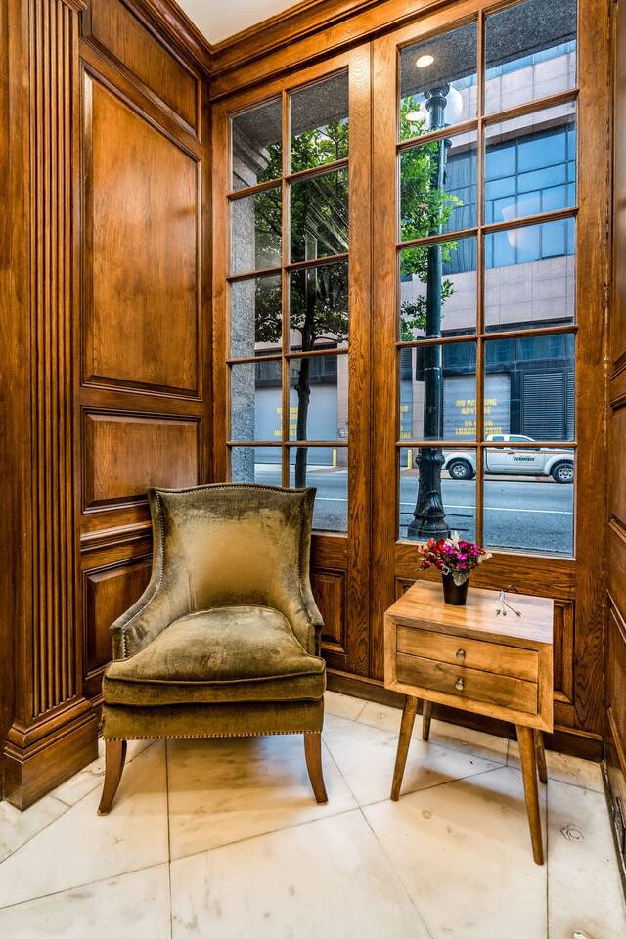 Une pièce d’angle avec un mur lambrissé en bois, un fauteuil confortable, une petite table d’appoint en bois sur laquelle est posé un vase de fleurs, et une grande fenêtre donnant sur la rue.
