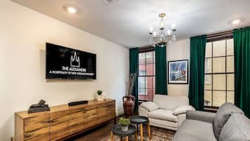A living room with a flat-screen TV, a wooden cabinet, a sofa, a coffee table, a chandelier, and a window with green curtains.