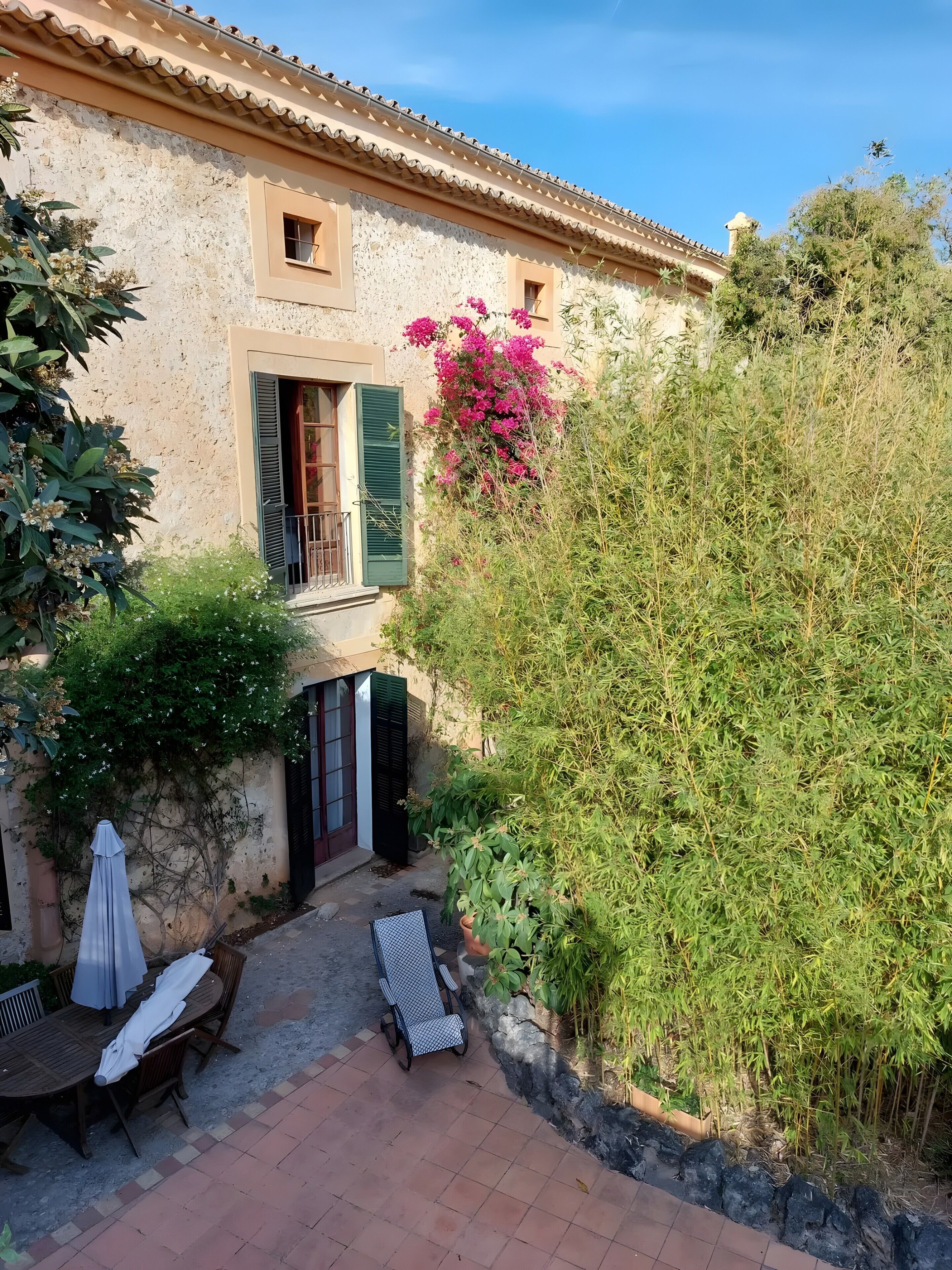 A courtyard with a table and chairs, a covered bench, a potted plant, and a building with a tiled roof.