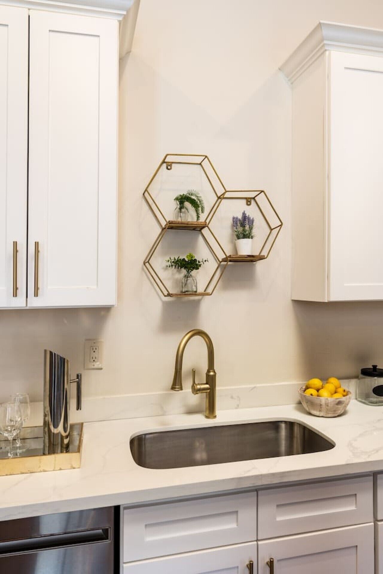 A kitchen with white cabinets, a stainless steel sink, and a gold faucet. There are three hexagonal shelves with plants and a bowl of lemons on the counter.