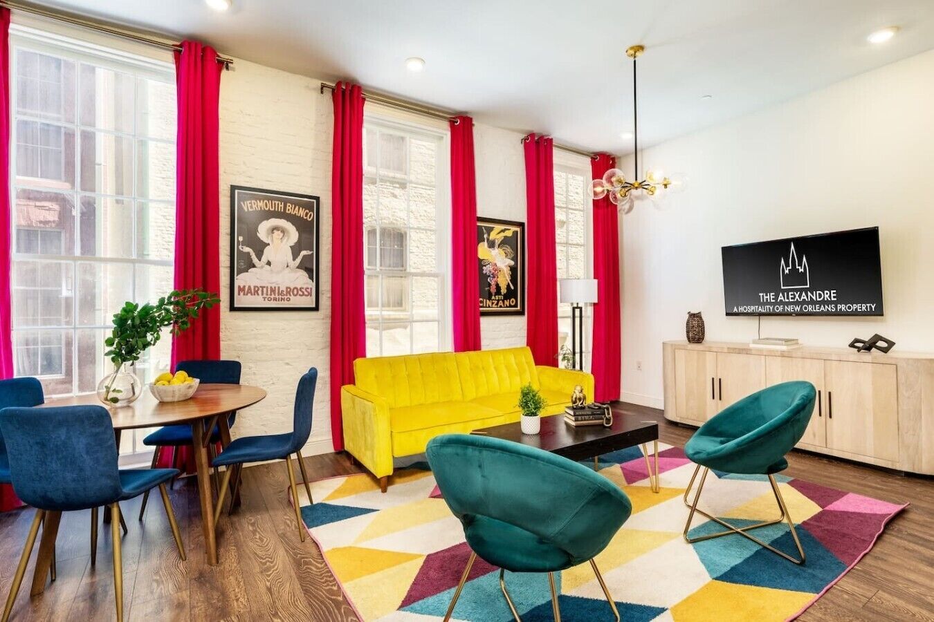 Een moderne woonkamer met een gele bank, blauwgroene stoelen, een houten eettafel met blauwe stoelen, rode gordijnen en een flatscreen televisie aan de muur.
