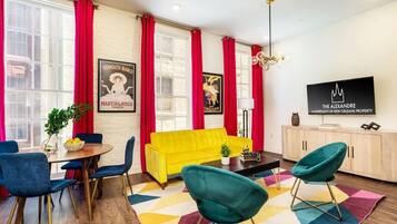 Een moderne woonkamer met een gele bank, blauwgroene stoelen, een houten eettafel met blauwe stoelen, rode gordijnen en een flatscreen televisie aan de muur.