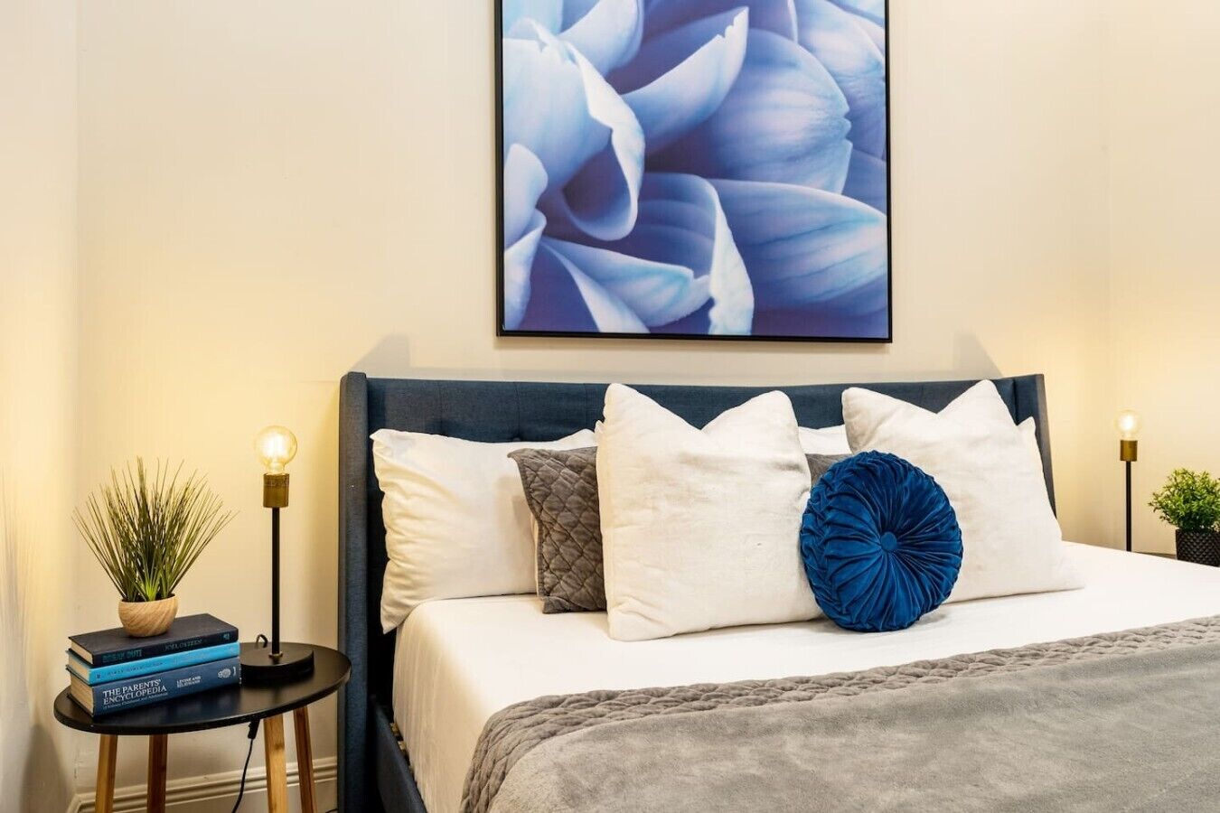 A neatly arranged bedroom with a bed, pillows, a bedside table with a plant and books, and a wall-mounted picture.
