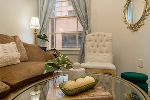 A living room with a brown sofa, a white tufted chair, a glass coffee table with decorative items, and a window with curtains. - Central 2BR Retreat | Bourbon & Streetcar Steps (New Orleans)