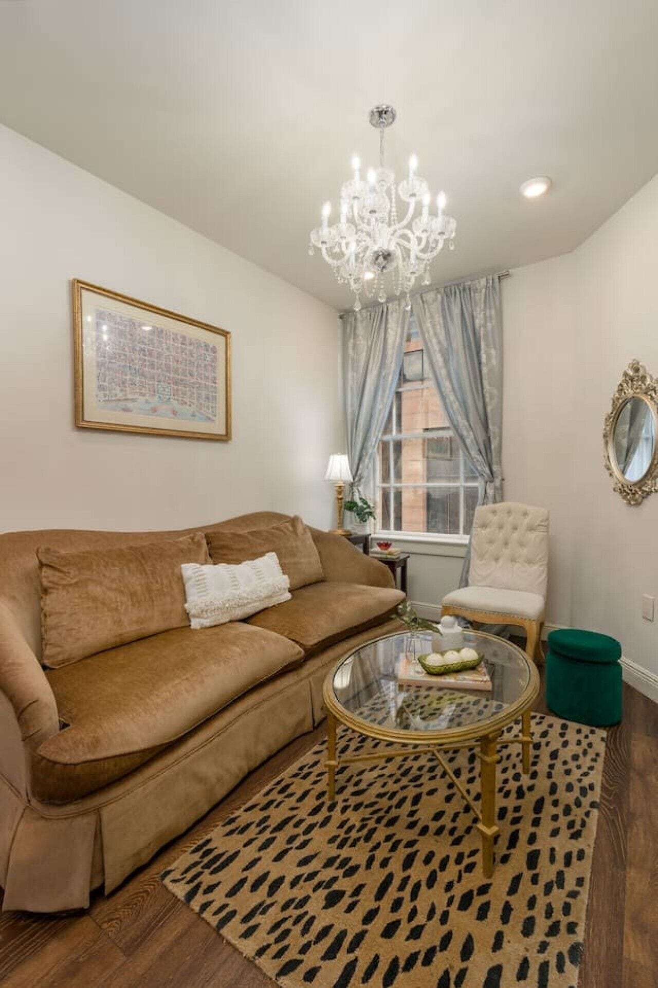 Un salon avec un canapé marron, une table basse en verre, un fauteuil à capitons, un lustre, un tableau encadré et une fenêtre avec des rideaux.