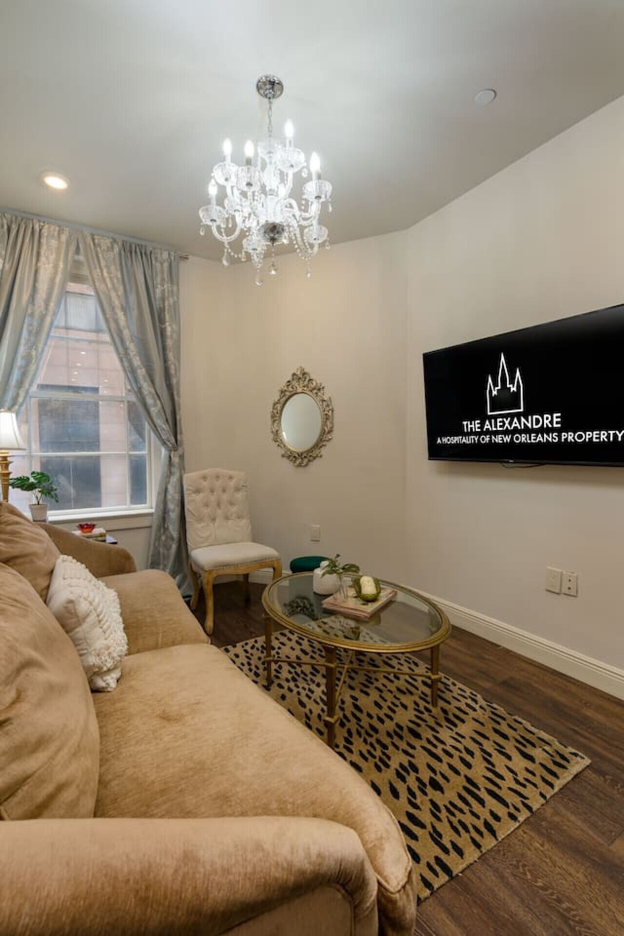 A living room with a beige sofa, a leopard print rug, a glass coffee table, a white armchair, a chandelier, a mirror, and a TV mounted on the wall.