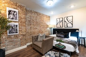 A hotel room with a brick wall, a bed with a dark headboard, a brown armchair, and two abstract wall art pieces. - Walk to Bourbon | Spacious & Bright 2BD in CBD (New Orleans)