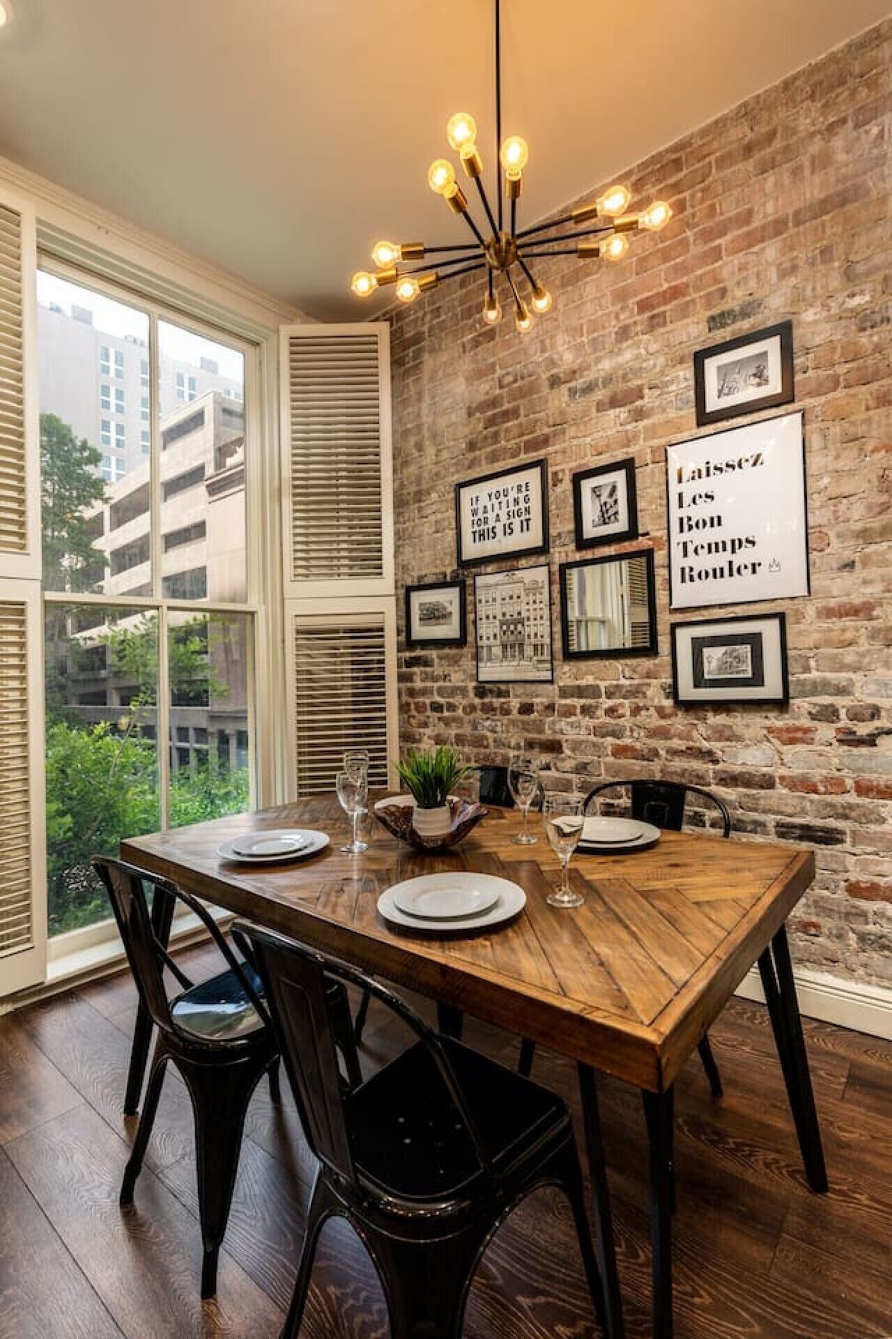 Un coin repas avec un mur en briques, des tableaux encadrés et une table en bois dressée pour quatre personnes.