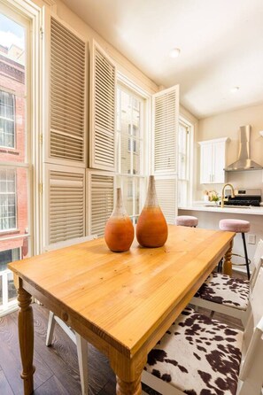 A wooden dining table with two vases, a kitchen area with white cabinets, and a view of a brick building through open shutters. - Group-Friendly NOLA Stay | Steps to French Quarter (New Orleans)