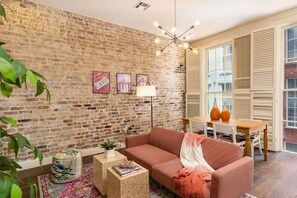 A living room with a brick wall, a pink sofa, a wooden coffee table, and a dining area with a table and chairs. - Group-Friendly NOLA Stay | Steps to French Quarter (New Orleans)