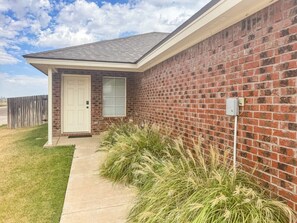 A residential building with a brick exterior, a white door, and a small porch area with grass and plants. - Pontiac Lubbock - 3-Bedroom Home in Lubbock, Texas (Lubbock)