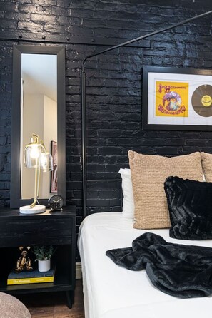 A bedroom with a black brick wall, a bed with white bedding, a black lamp, a framed picture, and a CD on the wall. - Prime NOLA Stay | 2BD Near Bourbon + Canal (New Orleans)