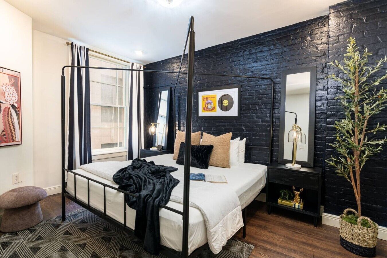 Una cama con dosel de estructura metálica negra, colchón blanco y manta negra. Hay una mesita de noche con una lámpara y una planta pequeña. La habitación tiene una pared de ladrillo negro y una ventana con cortinas.