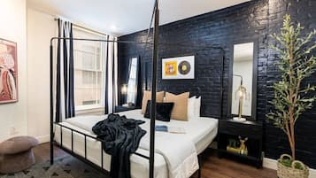 Una cama con dosel de estructura metálica negra, colchón blanco y manta negra. Hay una mesita de noche con una lámpara y una planta pequeña. La habitación tiene una pared de ladrillo negro y una ventana con cortinas.