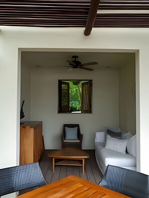 Une pièce avec un ventilateur de plafond, une fenêtre dont les volets sont ouverts, une table en bois, un canapé et un petit meuble.