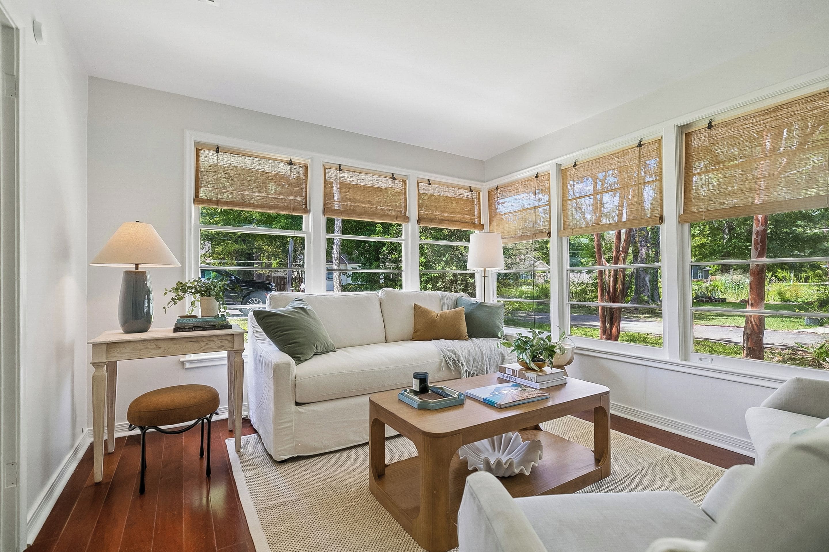A living room with a sofa, coffee table, and a side table with a lamp. Large windows with blinds are visible.