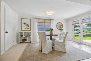 A room with a dining table, chairs, a rug, a shelf with decorative items, a framed picture, a window with curtains, and a view of greenery outside.
