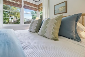 A bedroom with a bed, pillows, a window with blinds, and a painting on the wall.