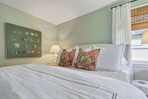 A bedroom with a bed, pillows, a nightstand, a lamp, a window with curtains, and a wall-mounted artwork.