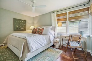 Ein Schlafzimmer mit einem Bett, einem Sessel, einem Schreibtisch, einem Fenster mit Jalousien und einem Deckenventilator.