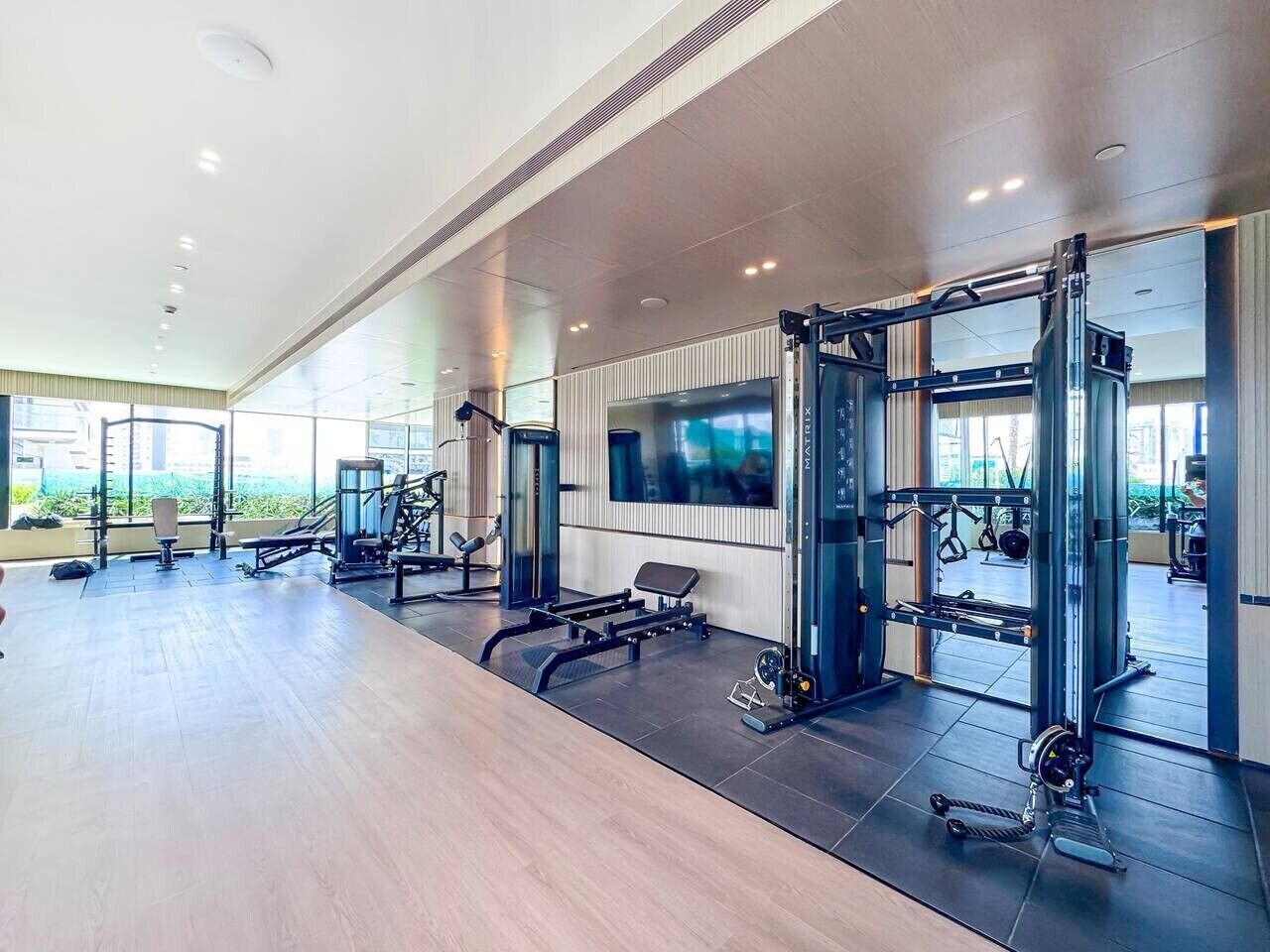 A gym with various exercise equipment, a television mounted on the wall, and a large window providing natural light.