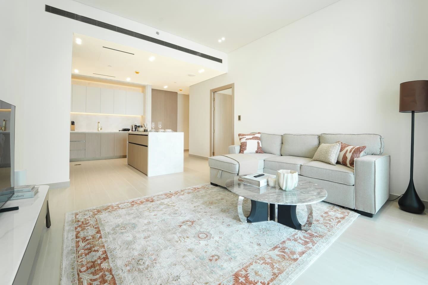 A modern living room with a sofa, coffee table, and a rug. There is a kitchen area visible in the background.