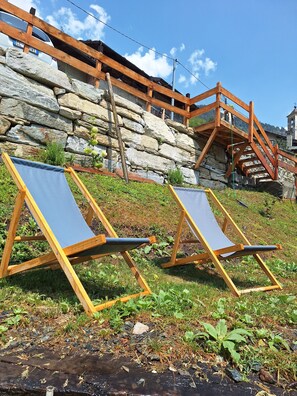 Zwei Holzliegen mit blauen Kissen auf einer grasbewachsenen Böschung, im Hintergrund eine Steinmauer und eine Holztreppe.