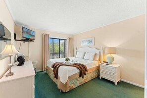 A hotel room with a bed, a desk, a chair, a television, a window with curtains, and a bedside lamp. - Tilghman 138- 3BR Ocean Drive Condo with Pool (North Myrtle Beach)