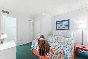 A hotel room with a bed, a desk, a chair, a lamp, a painting, and a closet. - Tilghman 138- 3BR Ocean Drive Condo with Pool (North Myrtle Beach)