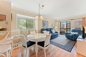 A spacious living area with a dining table, chairs, a sofa, and a balcony with a view. - Tilghman 138- 3BR Ocean Drive Condo with Pool (North Myrtle Beach)