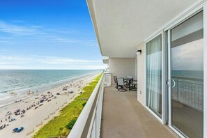 A balcony with a view of a beach and ocean, a table and chairs, and a glass door. - Oceanfront Crescent Shores 1204- 2 BR- Hot Tub (North Myrtle Beach)