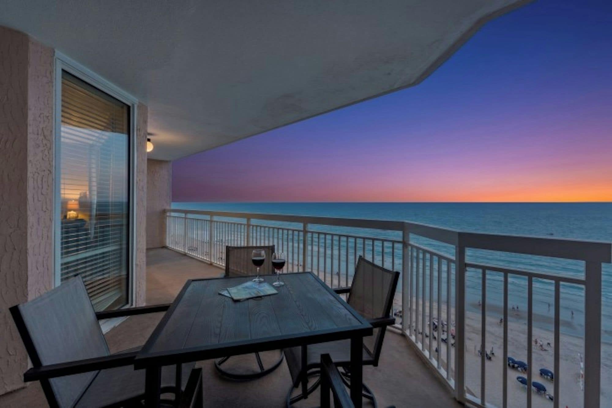 Un balcón con mesa y sillas, con vista a una playa al atardecer.