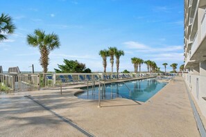 A pool area with a clear sky, palm trees, and a building with balconies. - Oceanfront Crescent Shores 1204- 2 BR- Hot Tub (North Myrtle Beach)