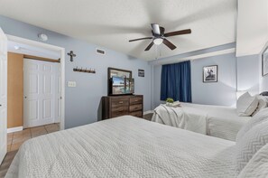 A bedroom with two beds, a ceiling fan, a dresser, a mirror, and a window with curtains. - Oceanfront 2BR Crescent Keyes 1103- I/O Pools (North Myrtle Beach)
