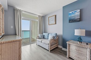 A room with a balcony, a wicker chair, a sofa with cushions, a side table with a lamp, and a framed artwork on the wall. - Oceanfront 2BR Crescent Keyes 1103- I/O Pools (North Myrtle Beach)