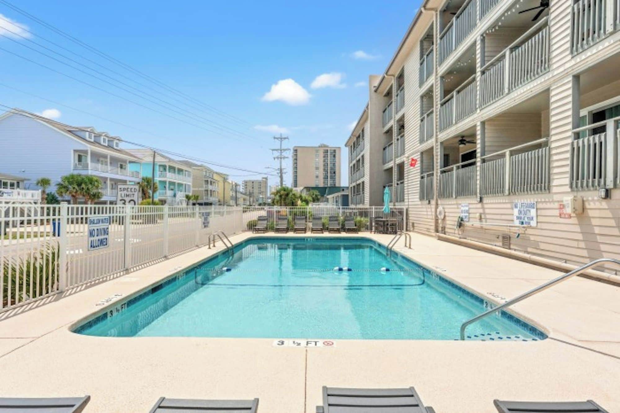 A swimming pool with a 3 1/2 ft depth marker, surrounded by a white fence, with a building featuring balconies and a lifeguard station in the background.