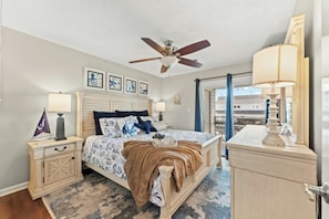 Une chambre à coucher avec un lit, des tables de chevet, un ventilateur de plafond, des œuvres d’art accrochées au mur et une vue sur l’extérieur par une porte ouverte.