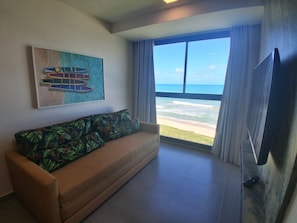 A living room with a sofa, a painting, a TV, and a large window with a view of the beach. - F1.302 | Exclusive Penthouse with Spa in Carneiros! (Tamandaré)