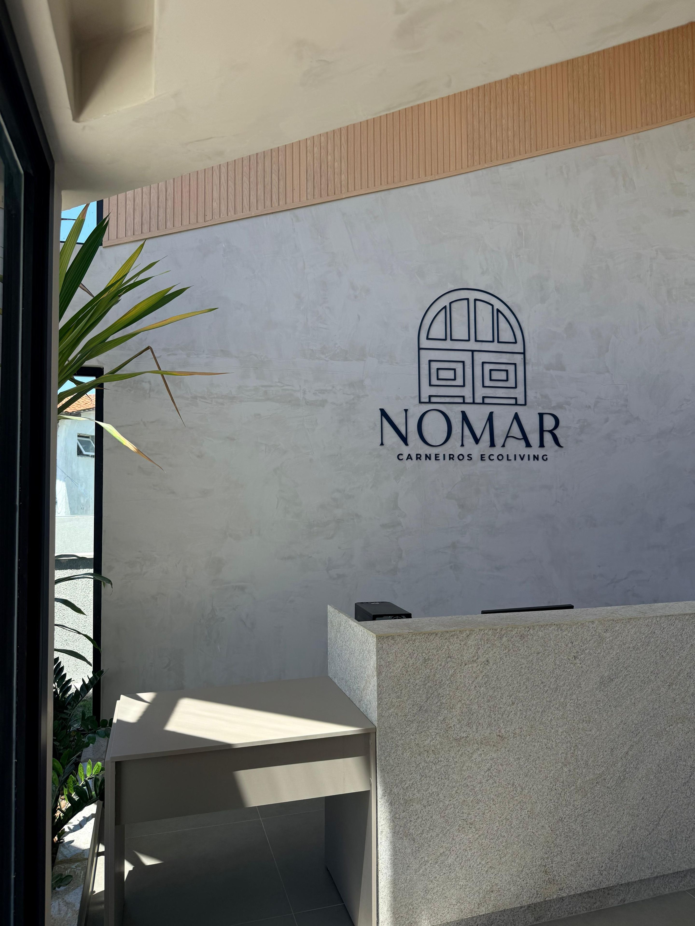 Balcão de recepção com o logotipo de 'NOMAR CARNEIROS ECOLIVING' em frente a uma parede texturizada.