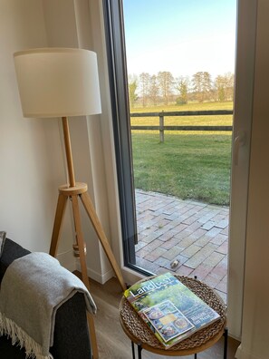 Una habitación con una lámpara de pie, un sillón de mimbre con una revista, y vista a un área verde a través de una puerta de vidrio.