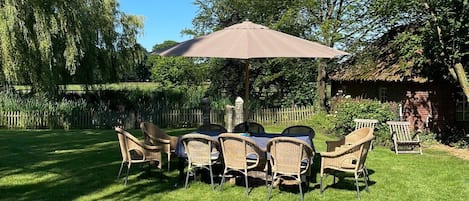 An outdoor seating area with a table and chairs under a large umbrella, surrounded by a well-maintained lawn and trees.