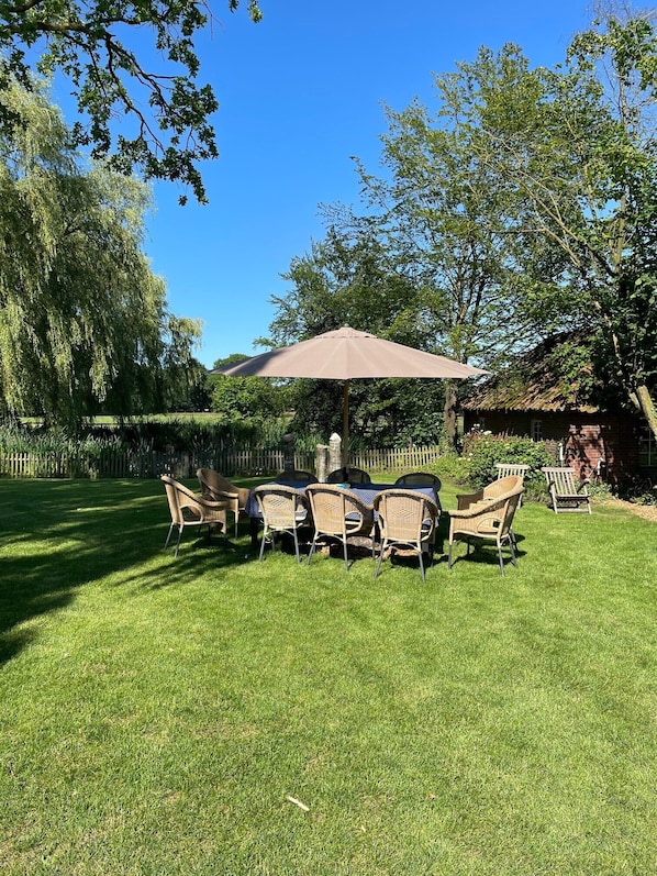 An outdoor seating area with a table and chairs under a large umbrella, surrounded by a well-maintained lawn and trees.